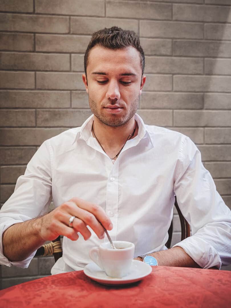 Ragazzo seduto a un bar davanti ad una tazza di caffè, vestito con camicia bianca a maniche risvoltate
